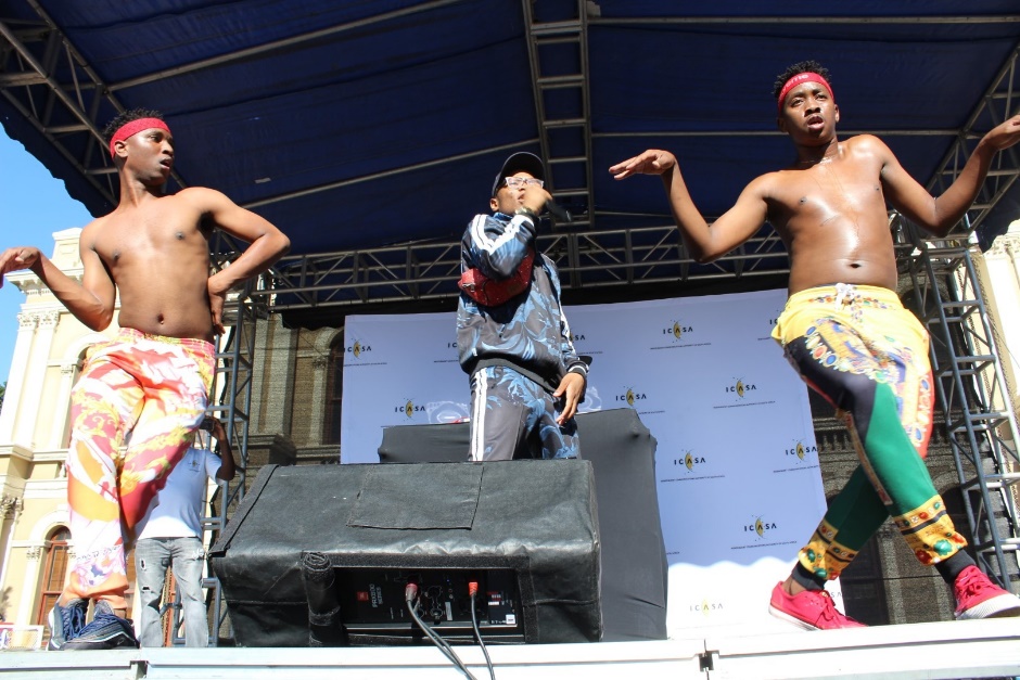 dancing and singing on stage at ICASA event; topless male performers wear colourful trousers